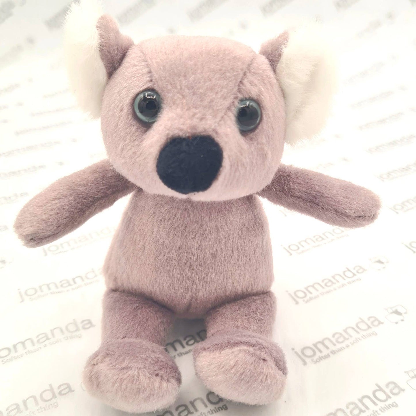 Plush koala toy with large eyes and a black nose on a white background with 'jomanda' branding.