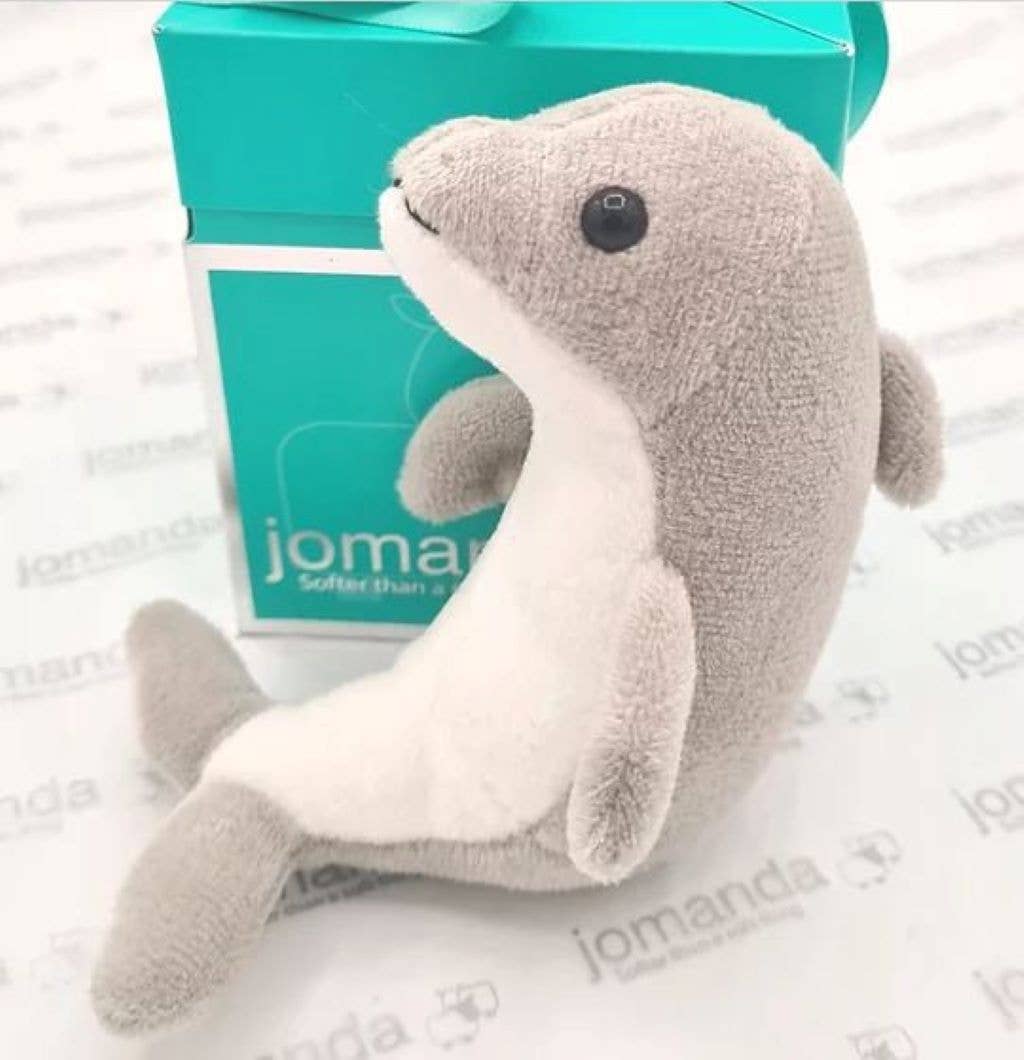 Plush dolphin toy in front of a teal box with 'jomanda' branding on a white background.
